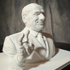 Può includere: Un busto bianco stampato in 3D di una persona in giacca e cravatta, raffigurato dal petto in su. La figura ha i capelli corti e una mano sollevata in un gesto. La superficie presenta linee di stampa orizzontali chiaramente visibili.