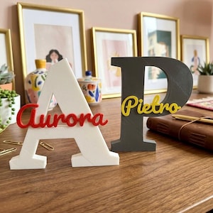 May include: Two freestanding 3D-printed letters, a white letter A with the red script name Aurora attached and a dark gray letter P with the yellow script name Pietro attached, displayed as decorative desk ornaments on a wooden surface.