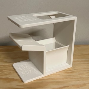 Op de afbeelding: Een witte plastic kaarten-schudmachine met een rechthoekige basis en een verhoogd bovenplatform. Het apparaat bevat reliëftekst op het bovenoppervlak "PLACE THE SHUFFLER ON THE TABLE" en op de basis "PLACE CARDS STRAIGHT INSIDE THE GUIDELINES."