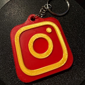 Può includere: Un portachiavi con il logo di Instagram rosso e oro. Il portachiavi ha una forma quadrata con angoli arrotondati, un contorno dorato e un cerchio dorato al centro. È attaccato a un portachiavi in metallo argentato.
