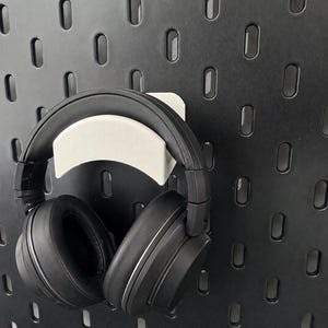 Könnte beinhalten: Ein schwarzer Over-Ear-Kopfhörer, der auf einer weißen, geschwungenen Hakenhalterung ruht. Die Halterung ist an einer schwarzen Lochplatte befestigt, die ein wiederkehrendes Muster aus vertikalen, ovalen Schlitzen zur Aufbewahrung aufweist.