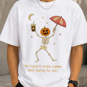 Pode incluir: Uma t-shirt branca de manga curta com um gráfico de um esqueleto com cabeça de abóbora segurando um café gelado e um guarda-chuva vermelho e amarelo sob uma lua crescente e estrelas, com o texto: me trying to enjoy summer while waiting for fall.