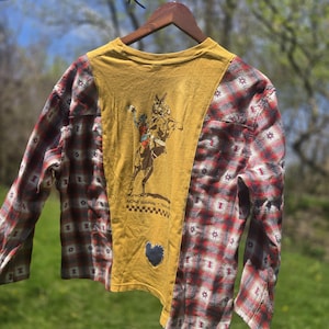 May include: Mustard yellow shirt with a cowboy graphic and a blue heart patch. The shirt has red and black plaid sleeves. The shirt is hanging on a wooden hanger against a blurred green background.