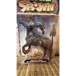 Op de afbeelding: Een Spawn Ultra actiefiguur in originele verpakking met een centaur-achtig wezen met donker pantser, paarse accenten en met bont bedekte hoeven. Het figuur houdt een hoge, puntige speer vast en bevat een klein, beige skeletachtig accessoire.