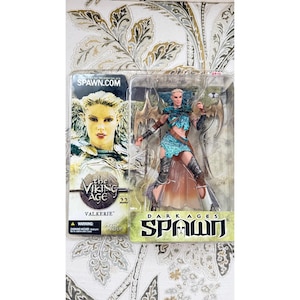 May include: A Spawn The Viking Age Valkerie action figure in its original plastic packaging. The figure features a blue and silver outfit with wings. The packaging includes the text SPAWN.COM, THE VIKING AGE, 22, VALKERIE, and SPAWN.