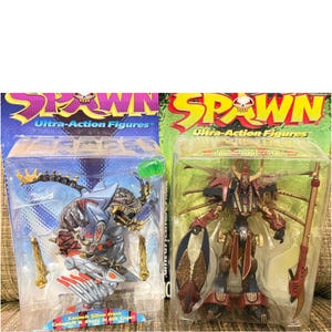 Könnte beinhalten: Zwei Spawn Ultra-Action-Figuren in Originalverpackung. Die linke Figur ist ein graues, haiartiges Wesen mit roten Akzenten und knochenartigem Zubehör. Die rechte Figur ist ein rot-goldener gepanzerter Krieger, der einen langen Stab und einen Schild hält.