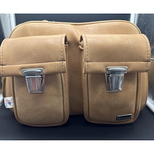 Peut inclure: Un sac en cuir beige doté de deux poches avant avec des fermetures à pression en métal argenté. Le sac comprend une petite étiquette sur le côté indiquant Made in USA et une plaque métallique rectangulaire sur la poche inférieure droite avec la marque Marsand.