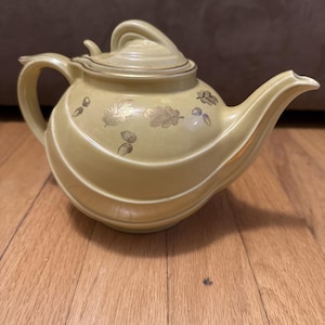 Peut inclure: Une théière en céramique jaune pâle dotée d'une anse incurvée, d'un couvercle assorti avec un bouton de préhension et d'un bec verseur. Le corps de la théière est orné d'un motif doré répétitif de feuilles de chêne et de glands.