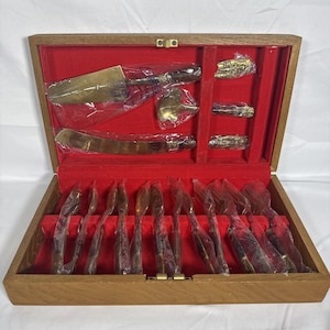 May include: A wooden storage box with a red velvet interior containing a set of gold-toned flatware and serving utensils. The pieces feature dark brown handles and are individually wrapped in clear plastic, including forks, knives, and spoons.
