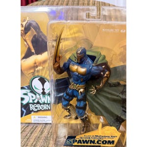 Peut inclure: Une figurine articulée McFarlane Toys Spawn Reborn dans son emballage plastique d'origine. La figurine porte une armure bleue et dorée, une cape verte et une grande lame incurvée sur le bras droit. L'emballage affiche le logo Spawn et du texte.