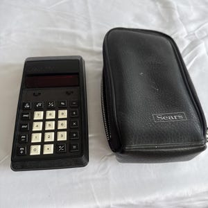Peut inclure: Une calculatrice électronique noire avec des touches numériques blanches et un écran sombre, placée à côté d'un étui de transport noir à fermeture éclair en matériau texturé avec le mot Sears imprimé en blanc sur le devant de l'étui.