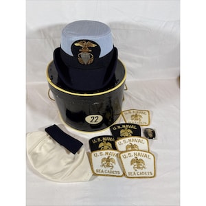 Puede incluir: Una gorra de estilo militar azul claro y azul marino oscuro con un emblema de águila dorada, sobre un contenedor de almacenamiento cilíndrico negro marcado con el número 22. Incluye una funda de tela blanca y varios parches bordados de U.S. Naval Sea Cadets.