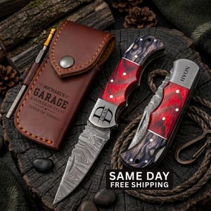 May include: A brown leather sheath with the text "MICHAEL'S GARAGE KNOWER OF STUFF FIXER OF THINGS" and a metal pen, alongside two folding knives with red and black marbled handles and Damascus steel blades, one engraved with "DEAN" and one with "NOAH".