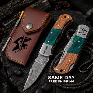 May include: A brown leather sheath with a metal snap and the name ROBERT printed above a climber graphic, alongside two folding knives featuring Damascus steel blades, wood and green resin handles, and the name HENRY engraved on the metal bolsters.