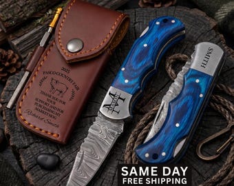 May include: Two folding knives with blue wood-grain handles and Damascus steel blades, one open and one closed. A brown leather sheath is engraved with '2025 PASCO COUNTRY FAIR' and 'Richard Smith'. A metal pen is tucked into the sheath side loop.