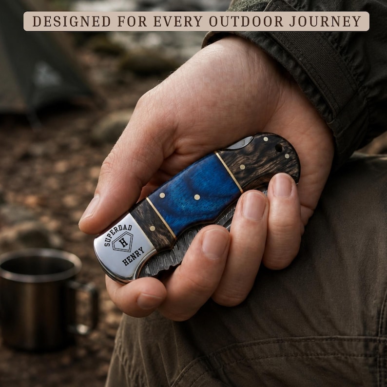 May include: A folding pocket knife with a dark wood and blue resin handle, featuring a silver bolster engraved with the text "SUPERDAD H HENRY". The blade has a textured finish, and the handle is secured with small brass pins and a lanyard hole.