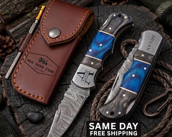 May include: Two folding pocket knives with Damascus steel blades and blue and black wood handles, one engraved with the name Adam and the other with Dylan, accompanied by a brown leather sheath stamped with Red Barn Farm Since 1985.