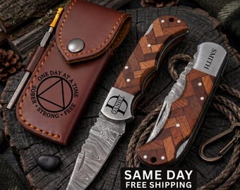 May include: A folding pocket knife with a Damascus steel blade and a herringbone wood handle, featuring the name DANIEL engraved on the bolster. Beside it, a closed knife with SMITH engraved, and a brown leather sheath with a sobriety symbol.