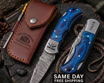 May include: A brown leather sheath with a metal snap and the text Out of Breath Hiking Society The King Family, alongside two folding knives with blue wood-grain handles, Damascus steel blades, and the word King engraved on the metal bolsters.