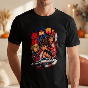 May include: A black t-shirt featuring a vibrant anime-style graphic print. The design displays three characters, a white sports car, and Japanese text. The artwork includes bold red, orange, and blue colors, with a sunset background and city silhouette.