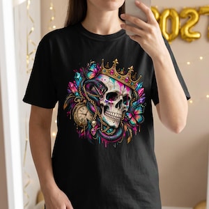 May include: A black t-shirt featuring a vibrant graphic print of a skull wearing a gold crown, entwined with a dark snake. The design includes colourful butterflies, crystals, a gold pocket watch, and a small skull charm with pink paint drips.