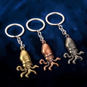 May include: Three metal octopus keychains in gold, copper, and silver tones. Each keychain features a smiling octopus charm attached to a chain and a keyring. The keychains are displayed on a dark blue background.