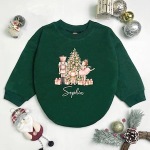 May include: A dark green long-sleeve sweatshirt featuring a central graphic of a nutcracker, a mouse, and a ballerina standing before a Christmas tree with gifts. The name Sophia is printed in white script below the festive holiday illustration.