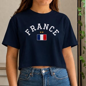 May include: Navy blue crop top with the word "FRANCE" in white arched lettering. Below the word is a small French flag and two gold stars. The shirt is paired with blue jeans.