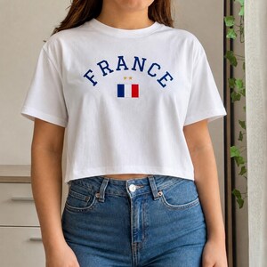 May include: White crop top with the word "FRANCE" in navy blue arched lettering. Below the word "FRANCE" is a small graphic of the French flag. The shirt is paired with blue jeans.
