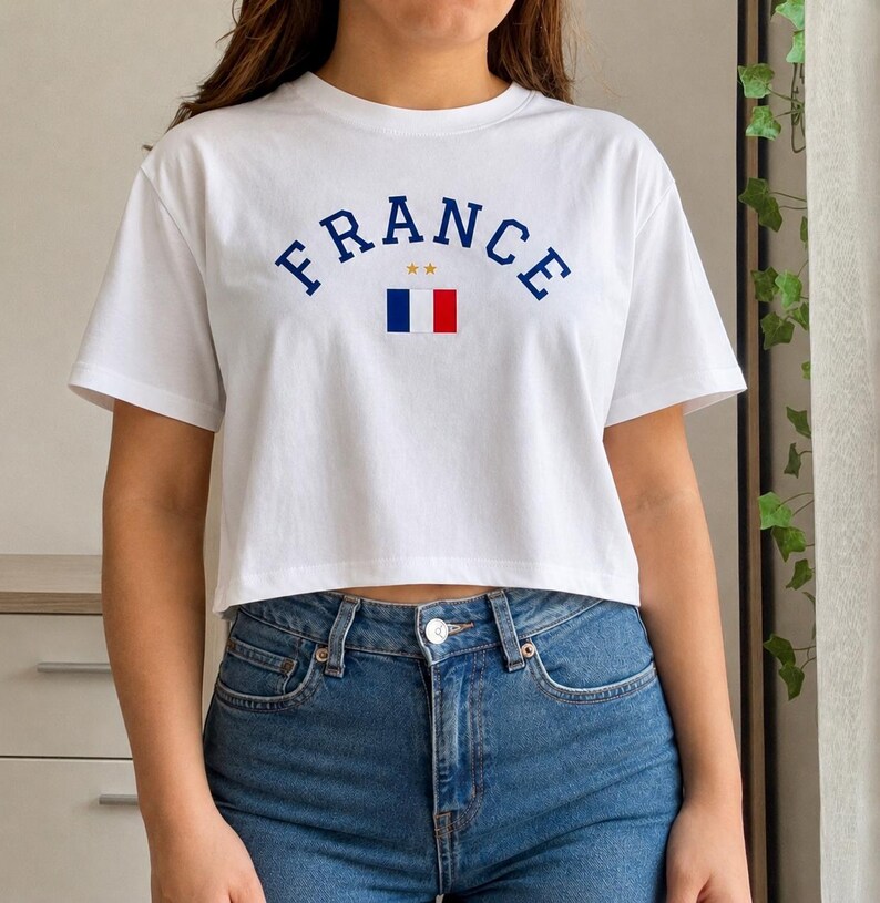 May include: White crop top with the word "FRANCE" in navy blue arched lettering. Below the word "FRANCE" is a small graphic of the French flag. The shirt is paired with blue jeans.