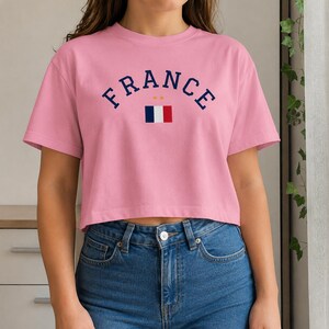 May include: A pink cropped t-shirt with the word "FRANCE" in navy blue arched lettering. Below the word "FRANCE" is a small French flag. The shirt is made of a soft material and is paired with blue jeans.