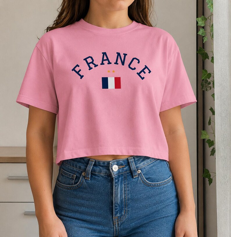 May include: A pink cropped t-shirt with the word "FRANCE" in navy blue arched lettering. Below the word "FRANCE" is a small French flag. The shirt is made of a soft material and is paired with blue jeans.