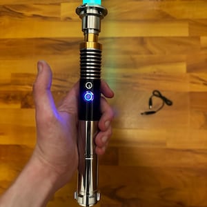 May include: A lightsaber with a glowing cyan blade and a metallic hilt featuring black and gold accents. The hilt includes a circular power button illuminated with a blue light. A small black charging cable is positioned separately to the side.