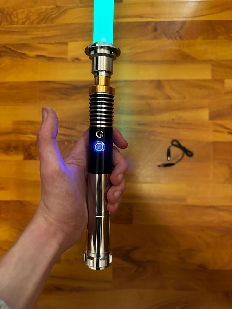 May include: A lightsaber with a glowing cyan blade and a metallic hilt featuring black and gold accents. The hilt includes a circular power button illuminated with a blue light. A small black charging cable is positioned separately to the side.