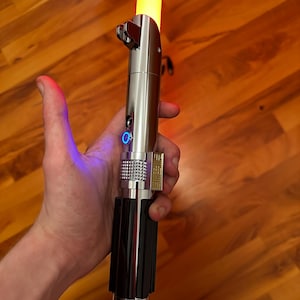 Könnte beinhalten: Ein silberner Lichtschwertgriff aus Metall mit einer leuchtenden orangefarbenen Klinge, die nach oben ragt. Der Griff verfügt über eine strukturierte Oberfläche mit schwarzen vertikalen Paneelen, einen blau beleuchteten Knopf und ein Steuergehäuse.