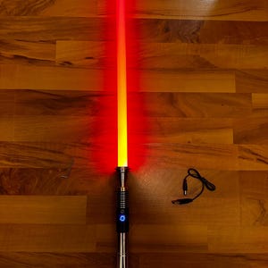 May include: A glowing red lightsaber with a metallic silver hilt featuring a circular power button with a blue light. The blade transitions from a bright yellow core to a vibrant red outer glow. A small L-shaped hex key and a black USB charging cable are included.