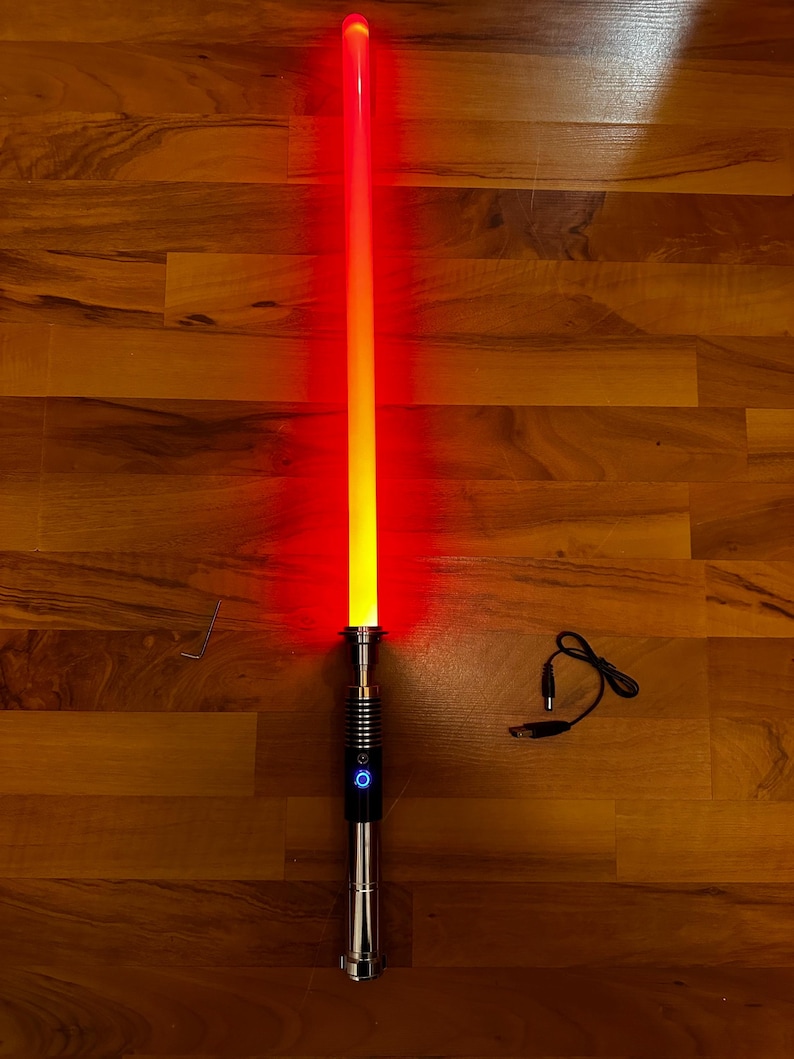 May include: A glowing red lightsaber with a metallic silver hilt featuring a circular power button with a blue light. The blade transitions from a bright yellow core to a vibrant red outer glow. A small L-shaped hex key and a black USB charging cable are included.