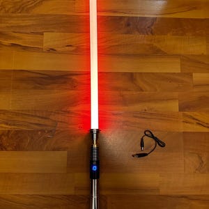May include: A glowing red light-up sword with a metallic silver and black handle featuring a circular blue power button. The set includes a small L-shaped metal tool and a black charging cable with a USB connector and a round power plug.
