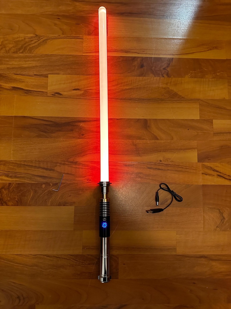 May include: A glowing red light-up sword with a metallic silver and black handle featuring a circular blue power button. The set includes a small L-shaped metal tool and a black charging cable with a USB connector and a round power plug.