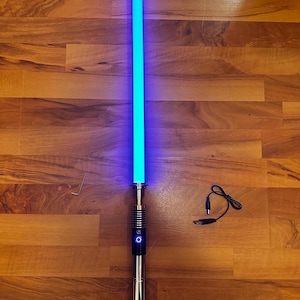 May include: A glowing blue light-up sword with a metallic silver and black handle featuring a circular power button. Included are a small L-shaped hex key and a black USB charging cable with a barrel connector for the hilt.