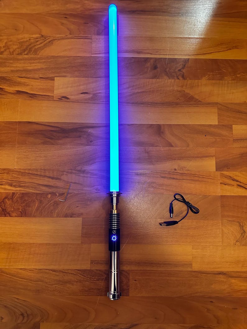 May include: A glowing blue light-up sword with a metallic silver and black handle featuring a circular power button. Included are a small L-shaped hex key and a black USB charging cable with a barrel connector for the hilt.