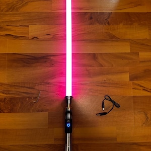 May include: A glowing pink lightsaber with a metallic silver hilt featuring a black grip section and a circular blue power button. The set includes a small L-shaped hex key and a black USB charging cable with a round connector plug.