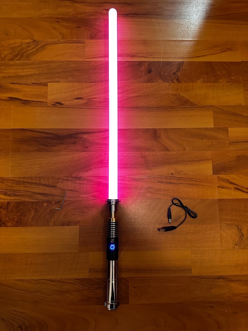 May include: A glowing pink lightsaber with a metallic silver hilt featuring a black grip section and a circular blue power button. The set includes a small L-shaped hex key and a black USB charging cable with a round connector plug.