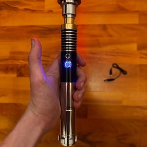 May include: A lightsaber with a polished silver and black metal hilt featuring a gold-colored neck section and a glowing yellow blade. The hilt includes a circular power button illuminated with a blue light ring and a textured grip area.
