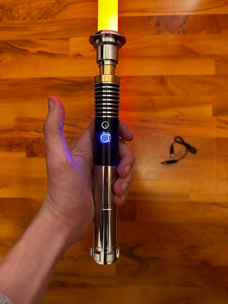 May include: A lightsaber with a polished silver and black metal hilt featuring a gold-colored neck section and a glowing yellow blade. The hilt includes a circular power button illuminated with a blue light ring and a textured grip area.