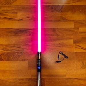 May include: A glowing pink lightsaber with a metallic silver hilt featuring a black grip section and a circular power button. The set includes a small L-shaped hex key and a black charging cable with a circular connector and a USB plug end.