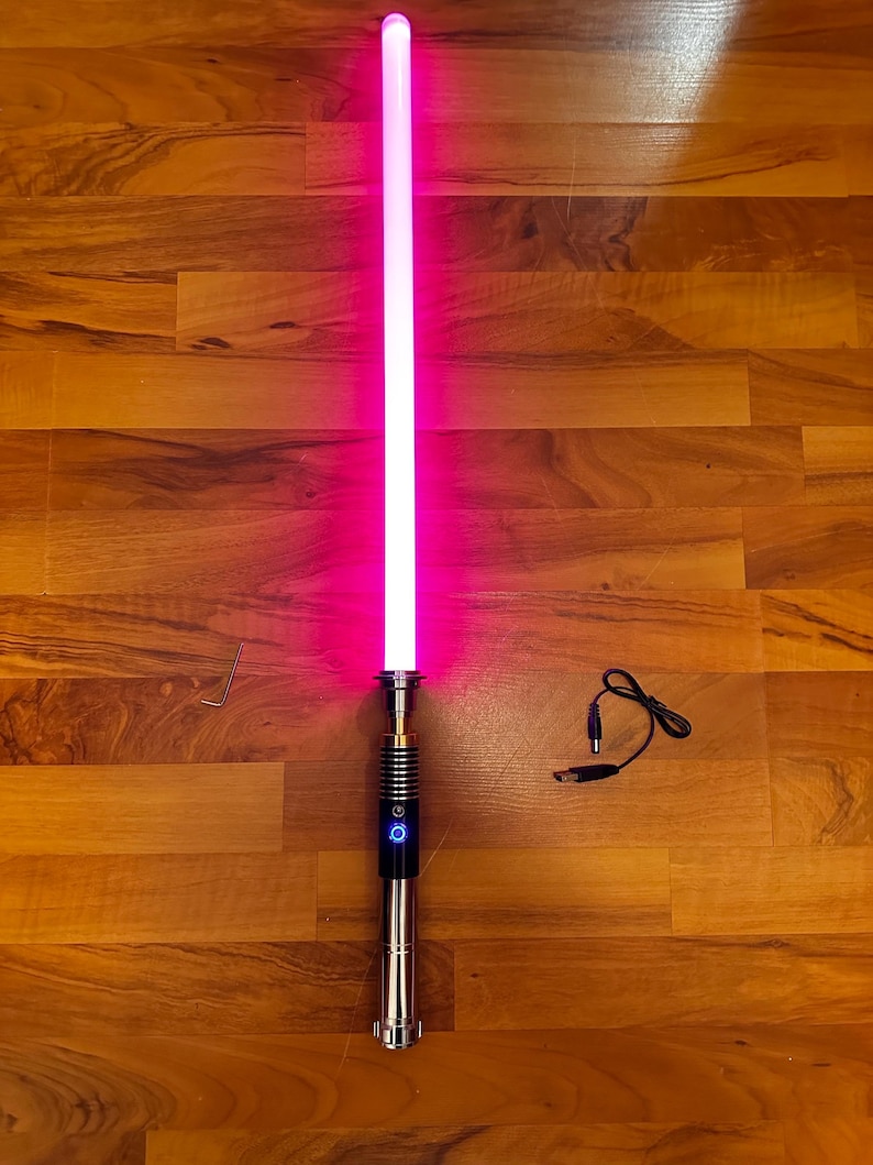 May include: A glowing pink lightsaber with a metallic silver hilt featuring a black grip section and a circular power button. The set includes a small L-shaped hex key and a black charging cable with a circular connector and a USB plug end.