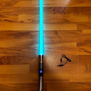 May include: A light-up toy sword with a glowing cyan blade and a metallic silver hilt featuring a circular power button. Included are a small L-shaped metal hex key and a black USB charging cable with a round barrel connector plug.