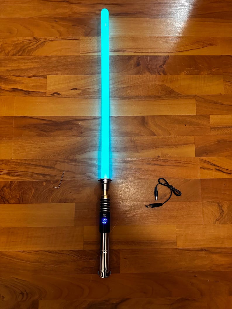 May include: A light-up toy sword with a glowing cyan blade and a metallic silver hilt featuring a circular power button. Included are a small L-shaped metal hex key and a black USB charging cable with a round barrel connector plug.