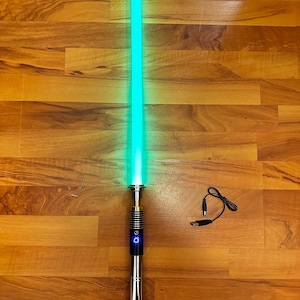 May include: A glowing green lightsaber with a metallic hilt featuring a black grip section and a blue illuminated power button. Included are a small L-shaped hex key and a black USB charging cable with a barrel plug connector for the device.
